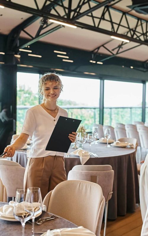 event-organizer-with-clipboard-pointing-at-table-with-festive-setting-near-woman-in-banquet-hall.jpg